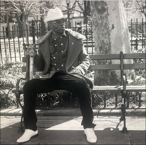Black-and-white portrait of LaRonz Murray seated on a park bench in a hat and tailored coat